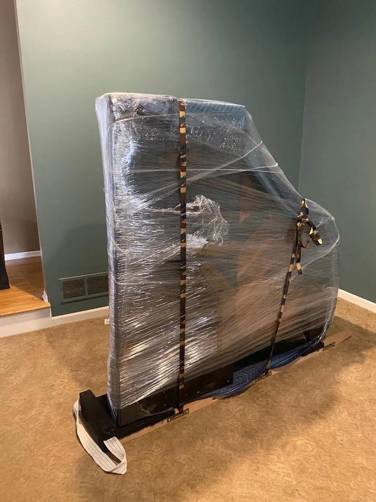 An upright piano wrapped in protective plastic and secured with straps on a dolly, ready for moving in a room with green walls.