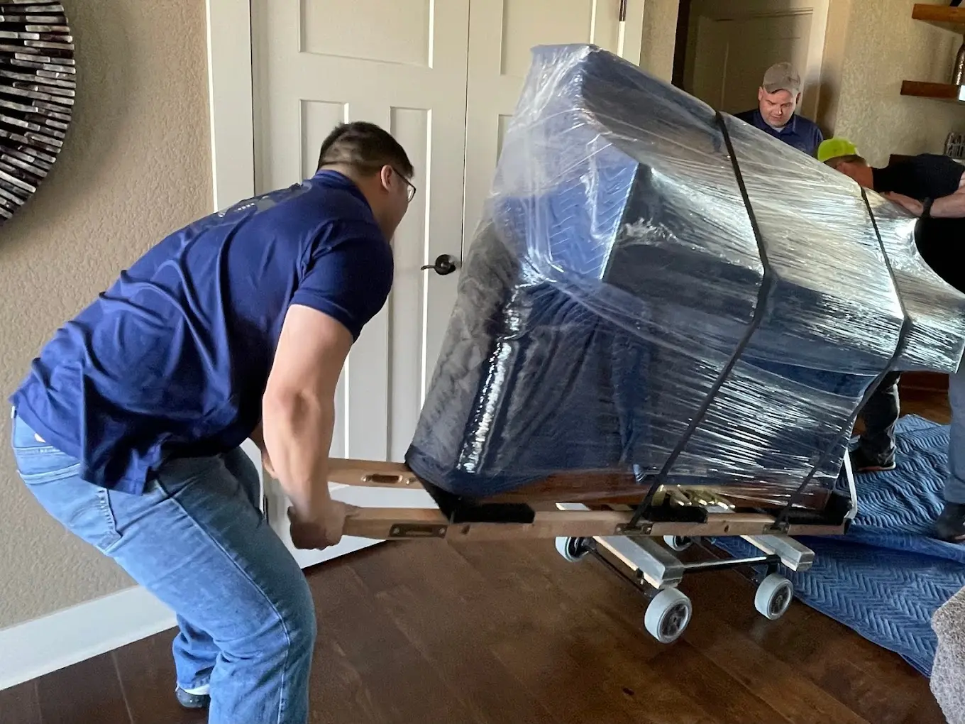 Two movers carefully maneuvering a large, plastic-wrapped piano on a dolly through a home, with a focused expression as they work to safely navigate the room.